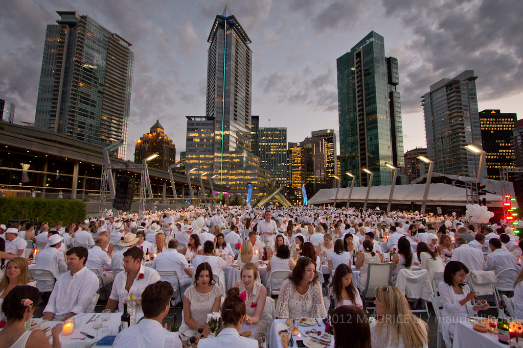 Hundreds of people dressed all in white dine outdoors at small tables crowded together.